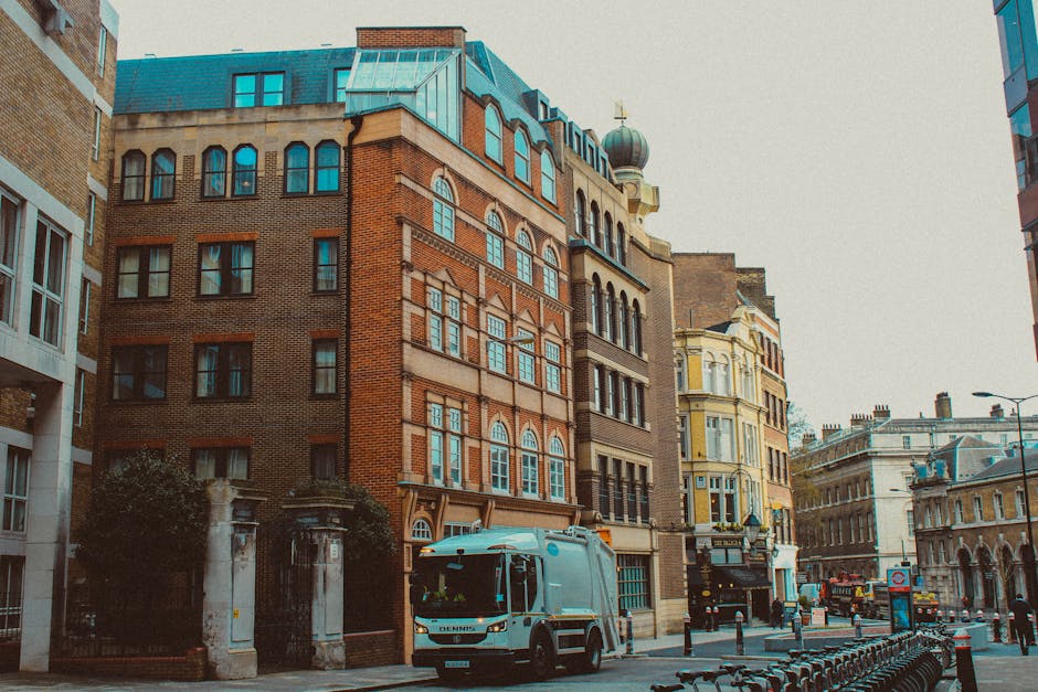A street scene featuring a light blue refuse collection truck parked in front of a row of historic multi-storey buildings with a mix of brick and stone facades. The truck, with a closed rear loading c
